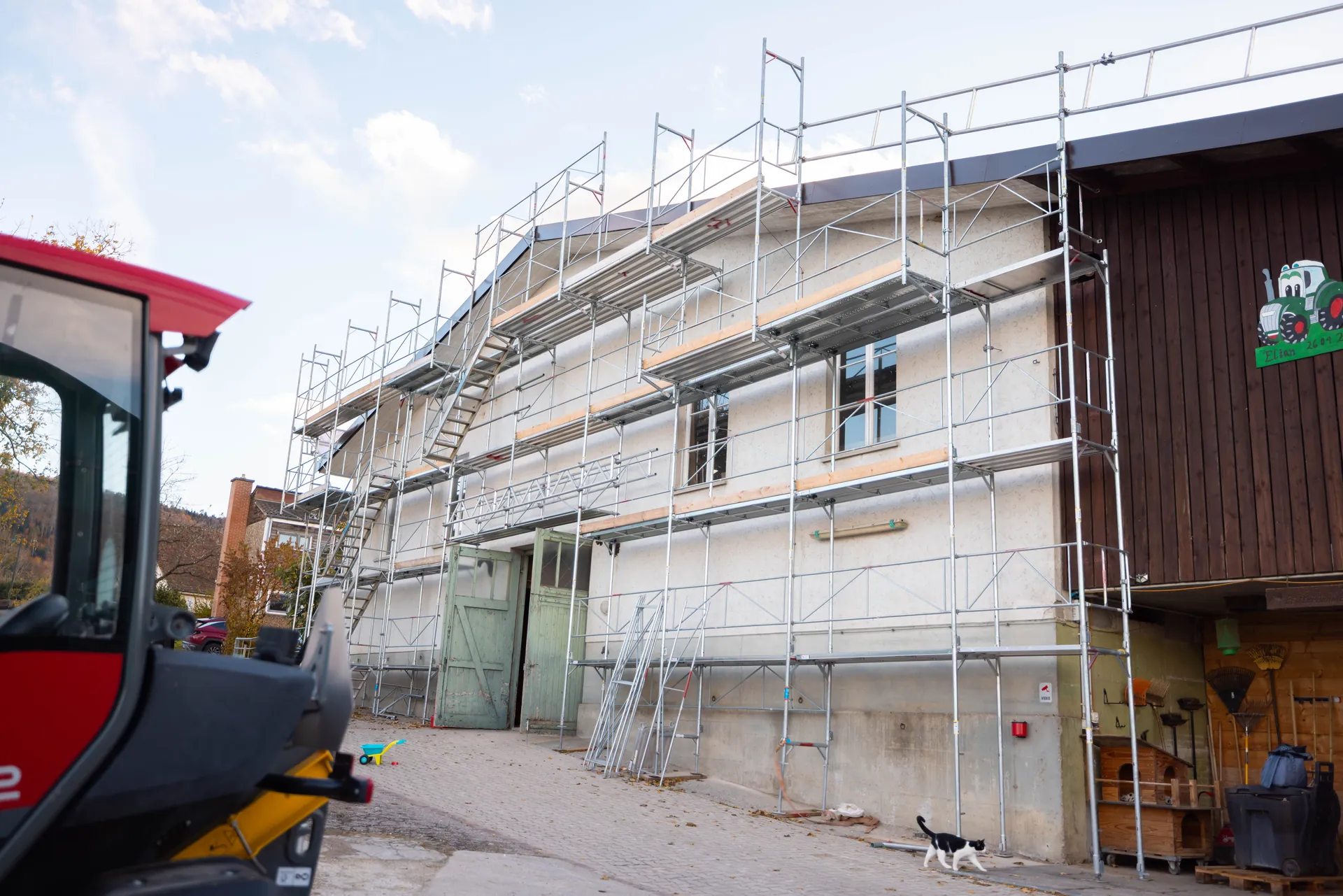 Lange Stallfassade, über die ganze Länge und Höhe eingerüstet, vorne angeschnitten eine Baumaschine