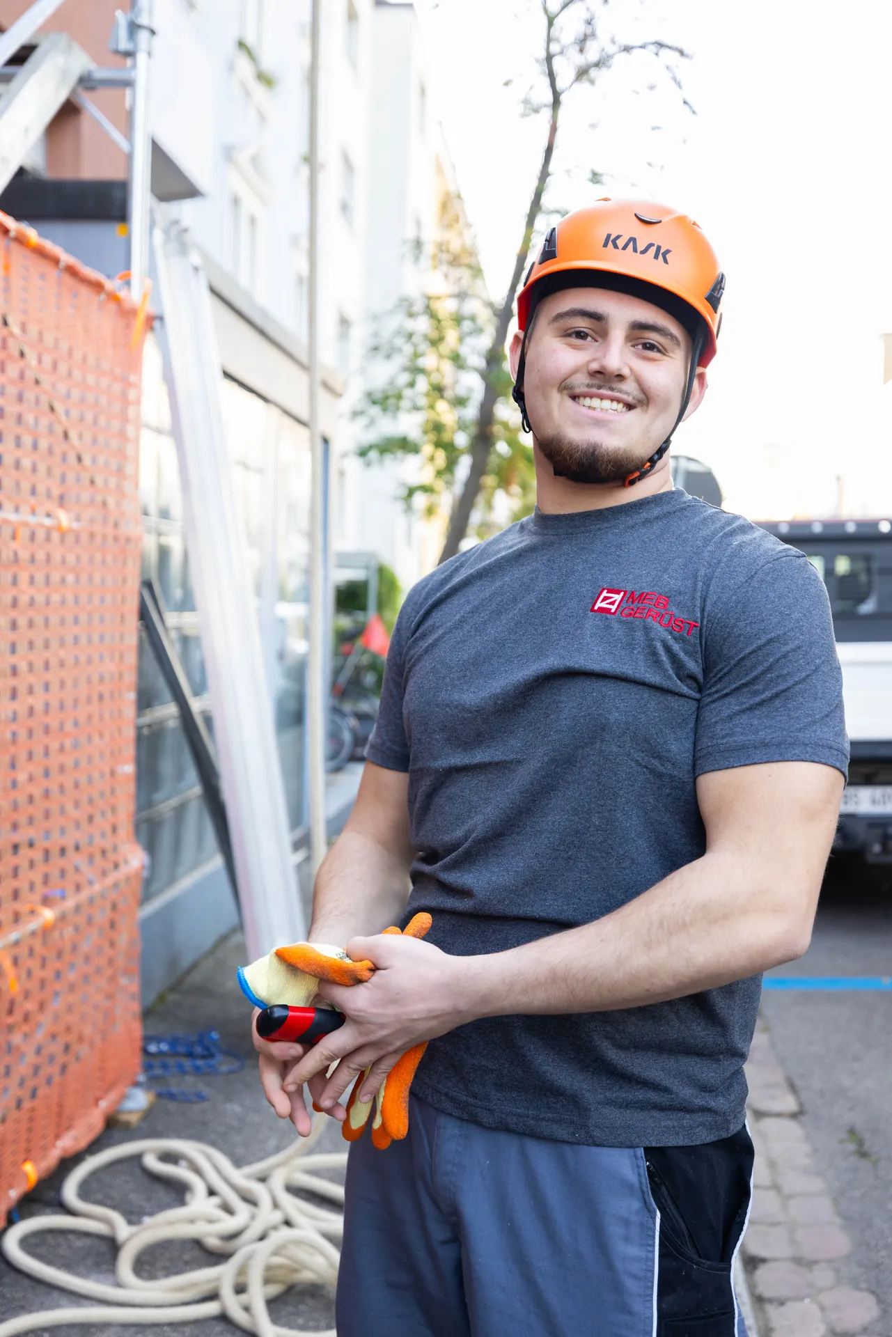 Junger Gerüstbauer im grauen MEB-Shirt mit orangem Helm lacht und hält seine Arbeitshandschuhe