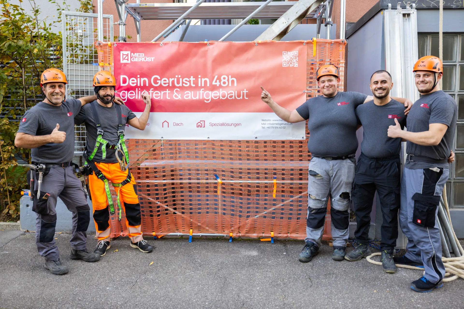 Fünf Gerüstbauer von MEB Gerüst zeigen lachend auf ihr Banner „Dein Gerüst in 48h geliefert & aufgebaut“