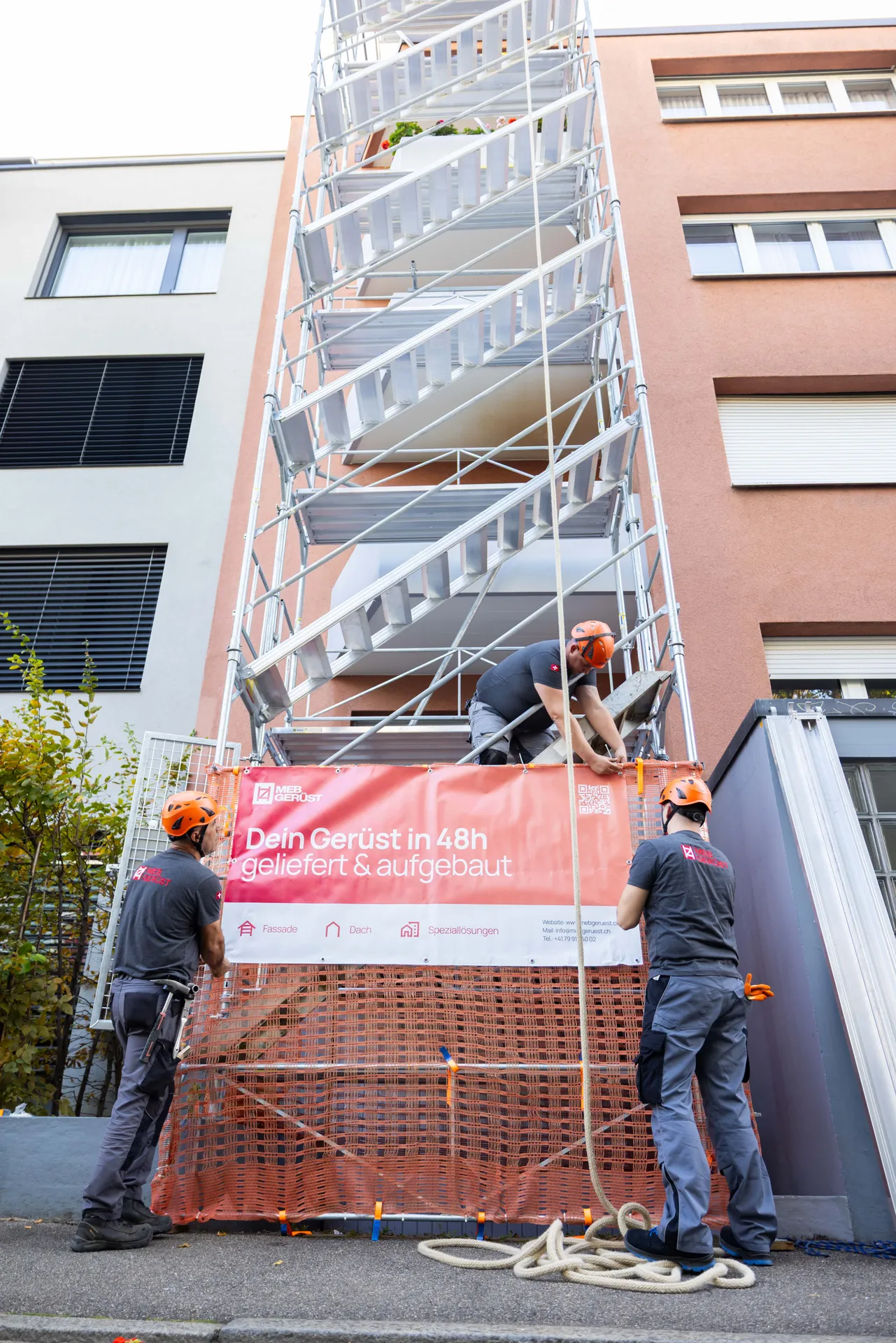 Treppenturm an einem Wohnhaus, unten hängen zwei Gerüstbauer das MEB-Banner auf
