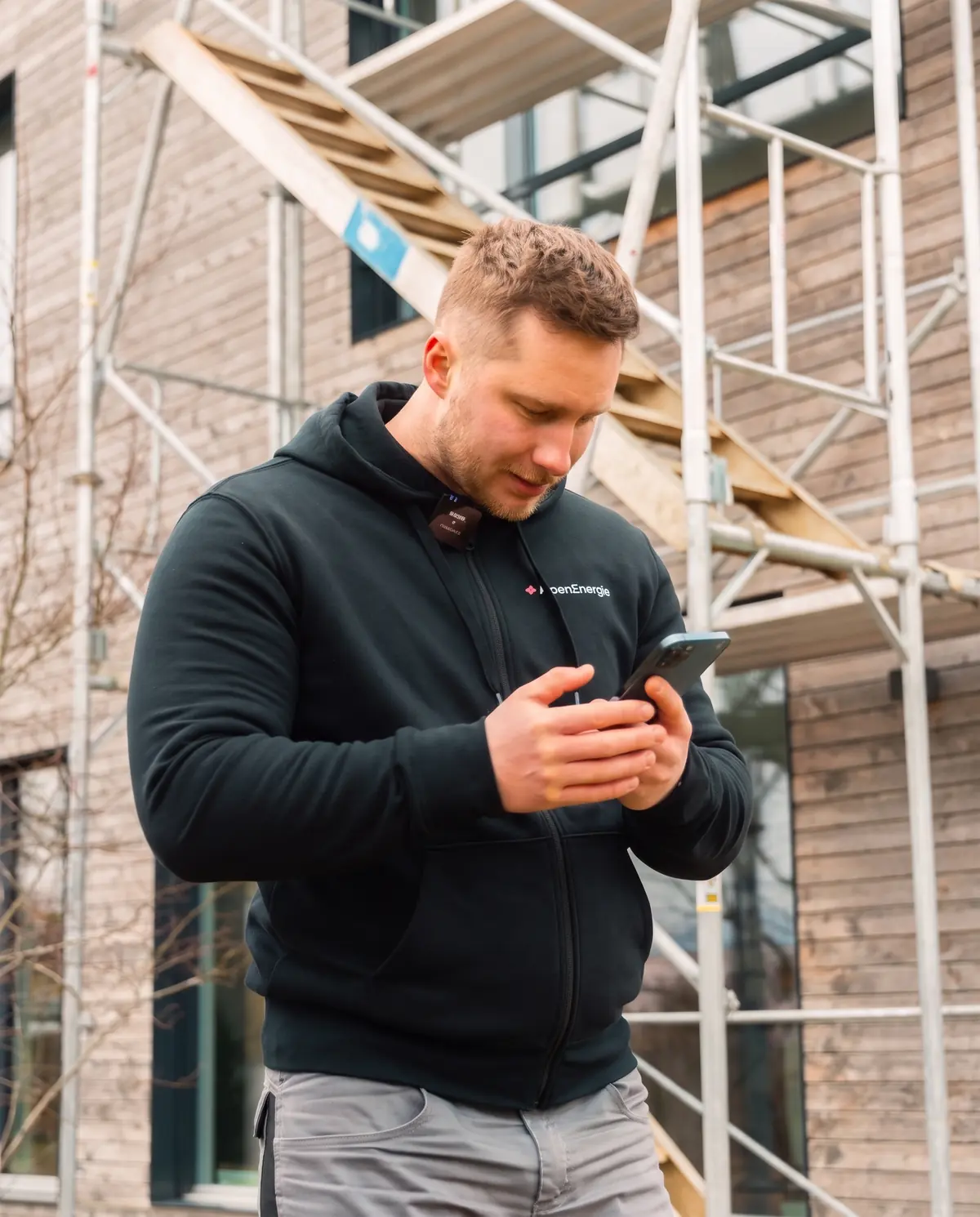 Mann im AlpenEnergie-Hoodie schaut vor einem Baugerüst auf sein Handy