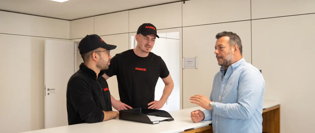 Zwei MAKE-Mitarbeiter im Gespräch mit einem Inhaber an dessen Empfangstresen