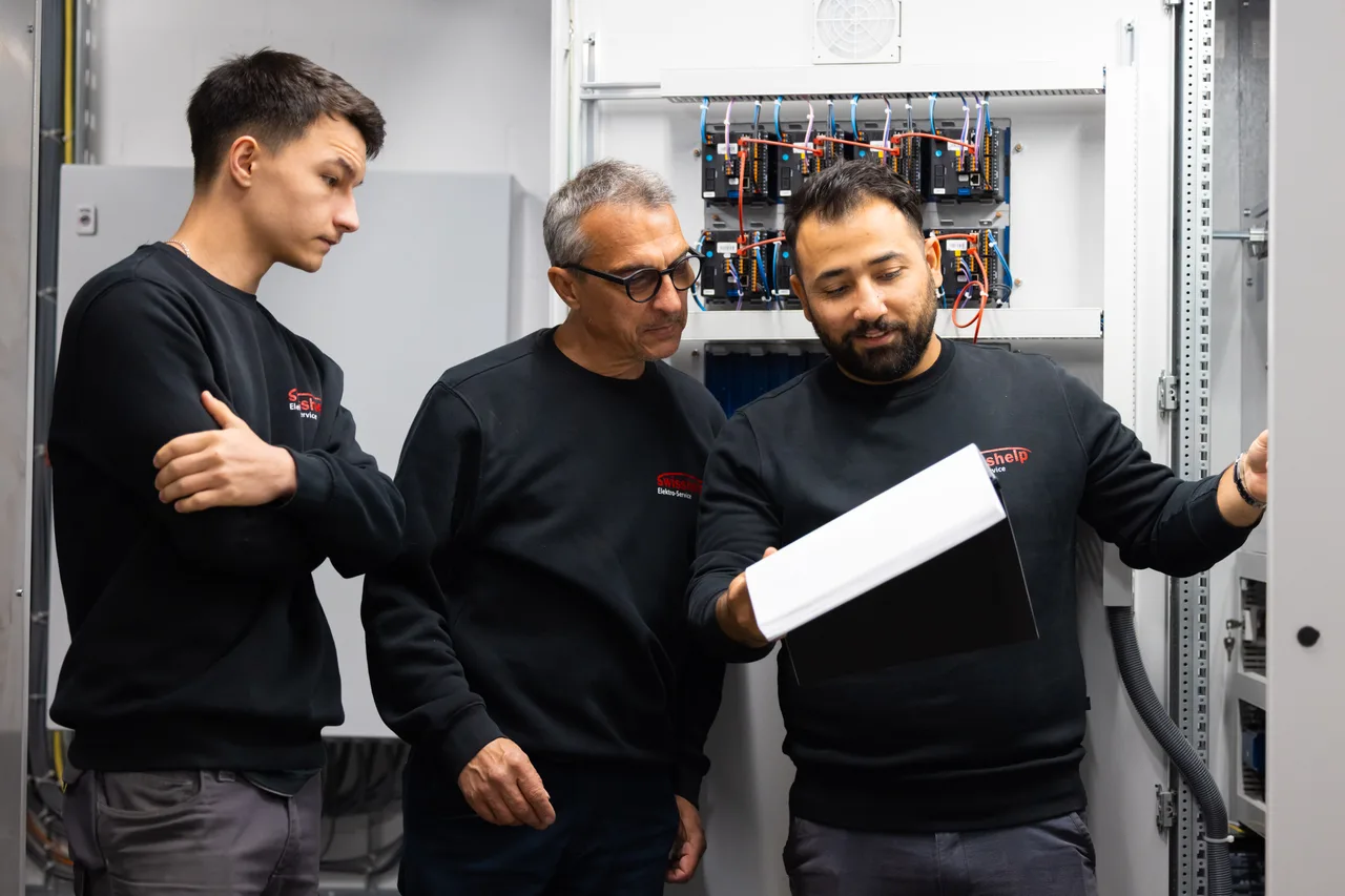 Drei Elektriker von Swisshelp besprechen vor einem offenen Schaltschrank einen Plan