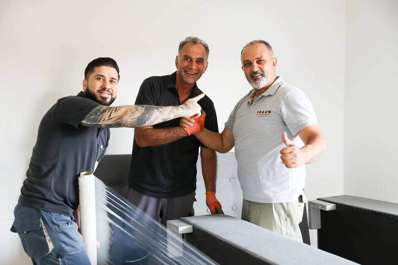 Drei Mitarbeiter lachen mit Daumen hoch beim Verpacken eines Betts