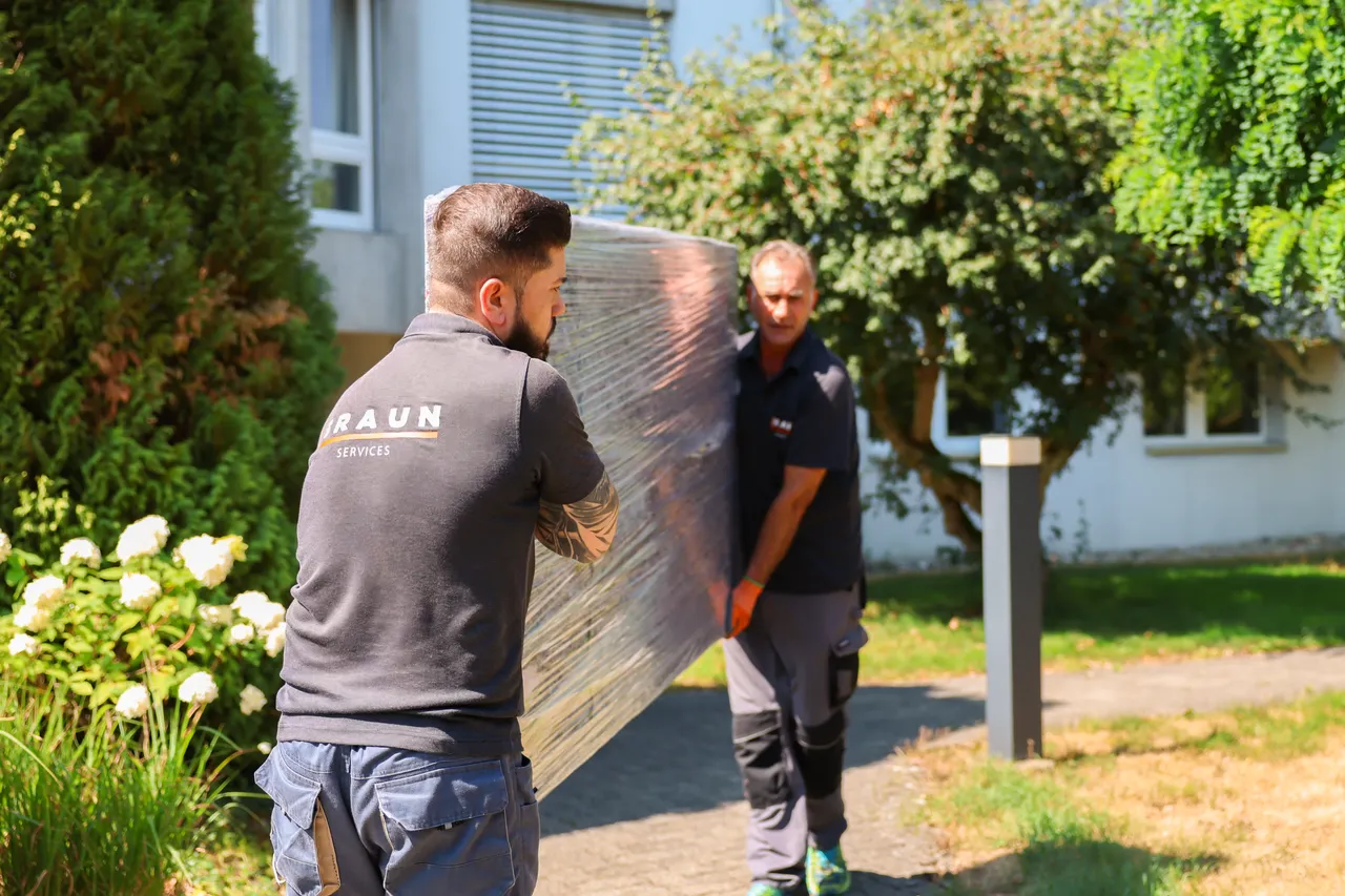 Zwei Mitarbeiter tragen ein verpacktes Möbelteil über den Weg vor einem Wohnhaus