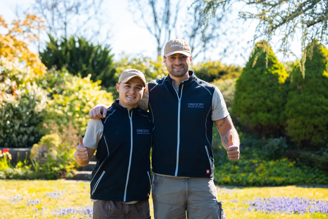 Gregory Wurmser und ein Mitarbeiter Arm in Arm mit Daumen hoch in einem blühenden Garten