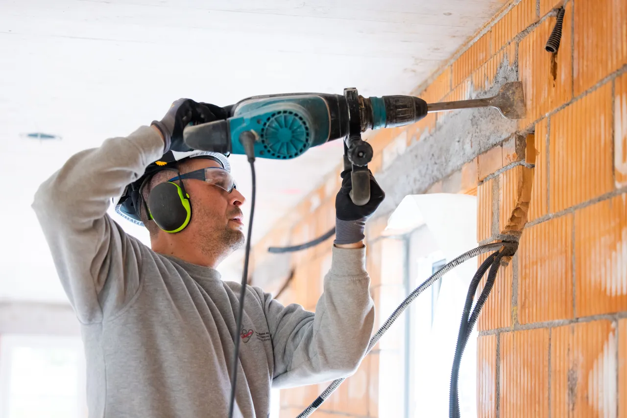 Elektriker mit Helm und Gehörschutz stemmt mit dem Bohrhammer an einer Backsteinwand im Rohbau