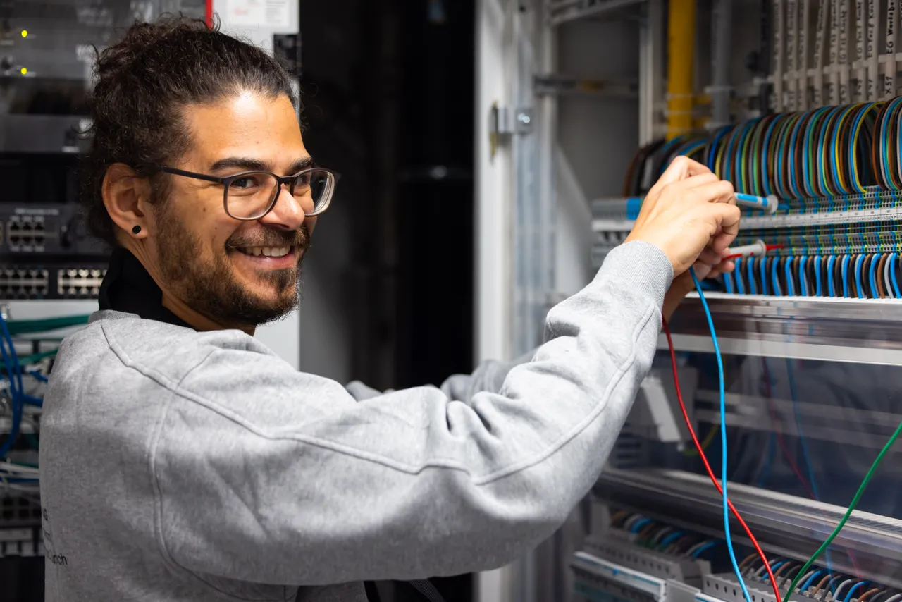 Elektriker mit Brille lacht in die Kamera und misst an Klemmen im Schaltschrank