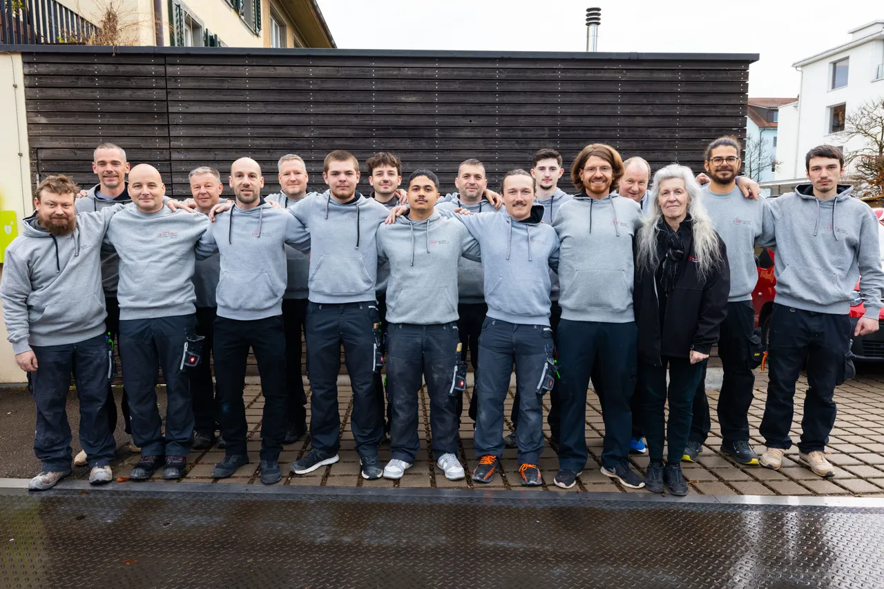 Das ganze Team von Heeb + Enzler in grauen Firmenpullis Arm in Arm vor einer dunklen Holzlattenwand