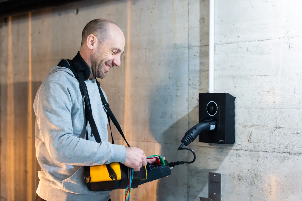 Elektriker prüft lachend eine Wallbox in einer Tiefgarage mit einem gelben Installationstester