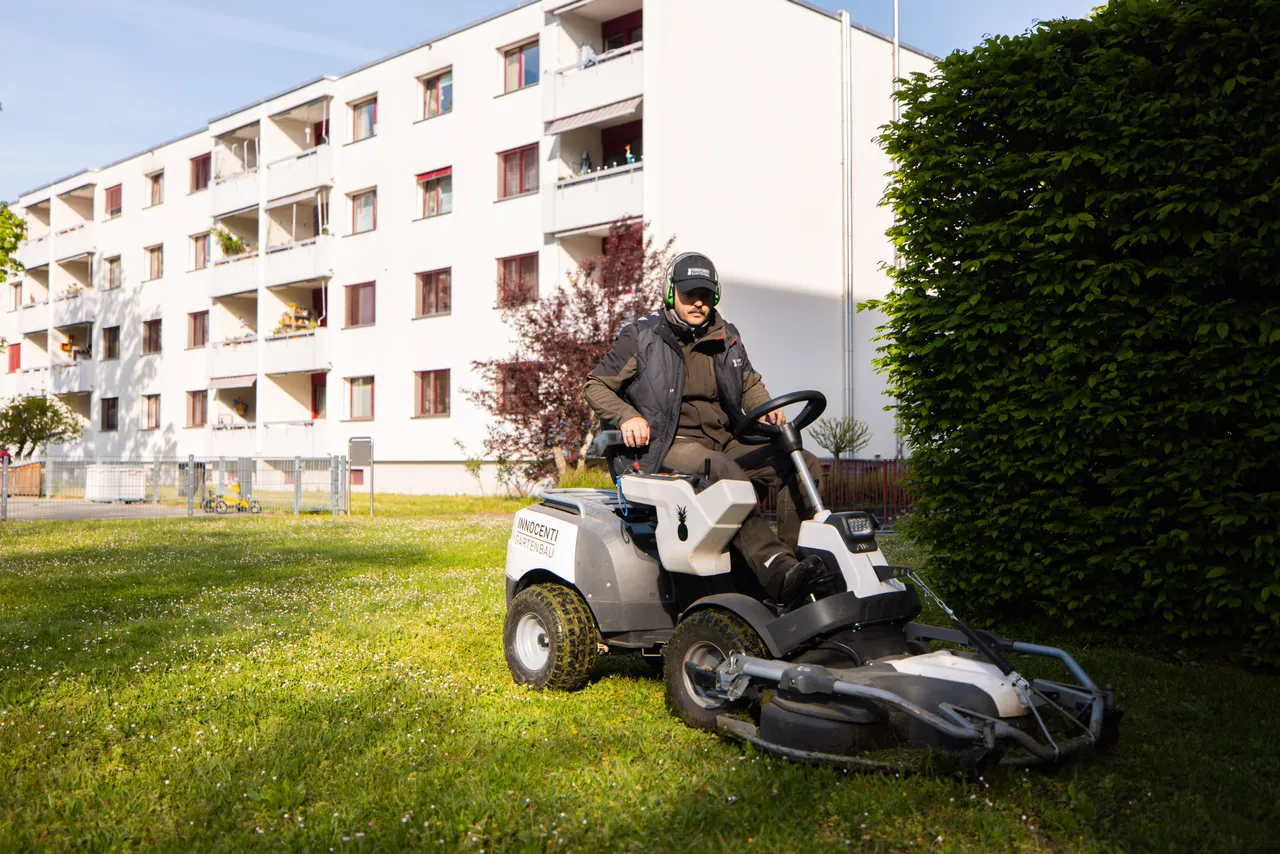 Gärtner auf dem Aufsitzmäher von Innocenti vor einem Wohnblock und einer Hecke