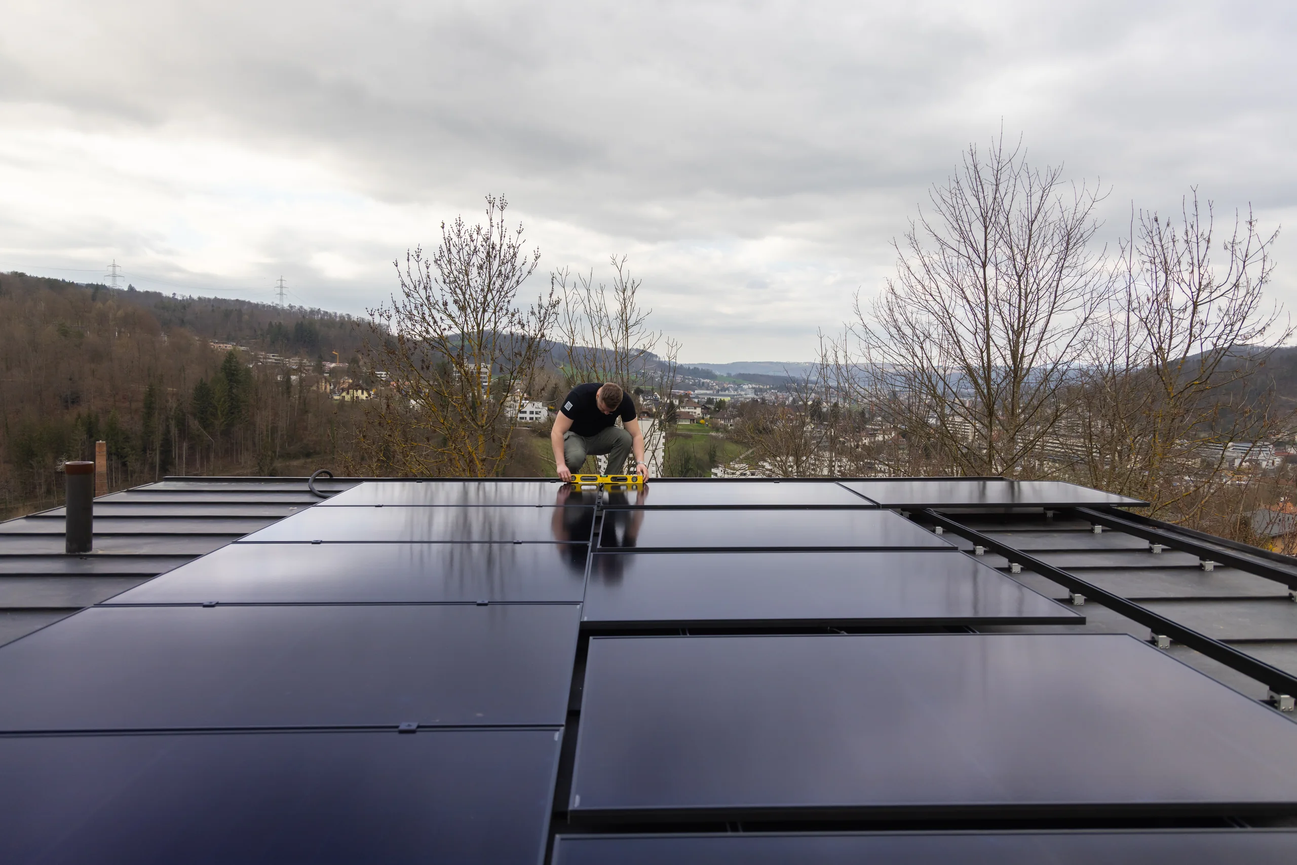Monteur richtet schwarze Solarmodule auf einem Flachdach aus, im Hintergrund Hügel und Siedlung