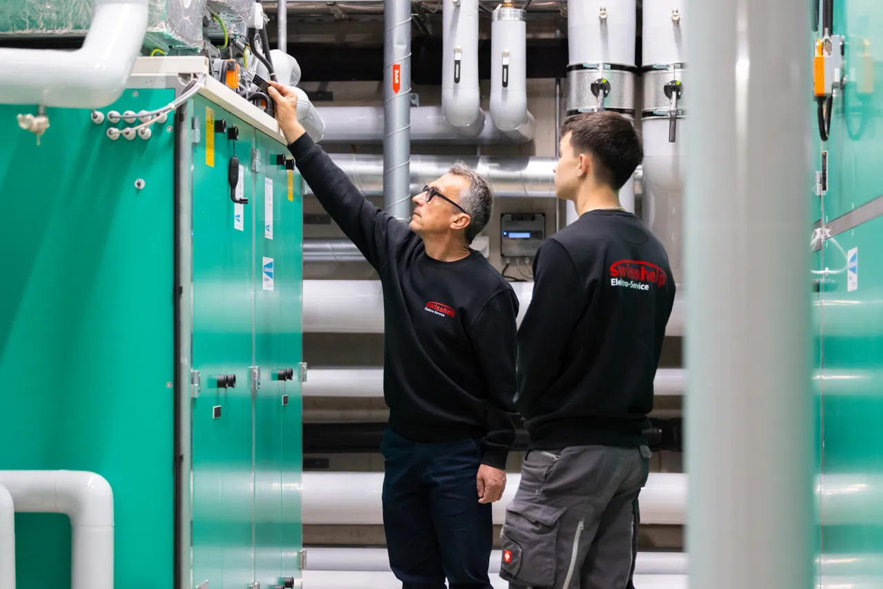 Zwei Elektriker prüfen die Steuerung einer grünen Lüftungsanlage im Technikraum