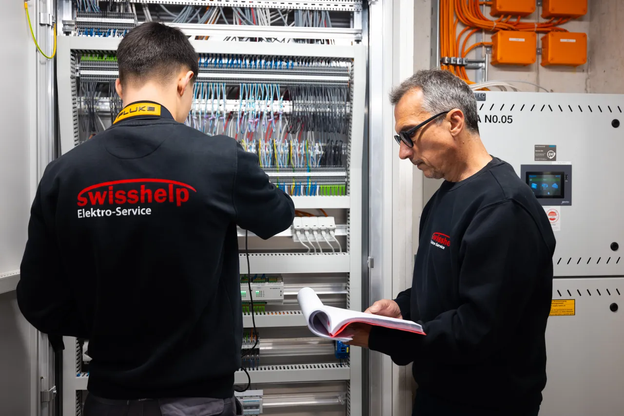 Junger Elektriker am offenen Schaltschrank, ein älterer Kollege liest daneben den Plan