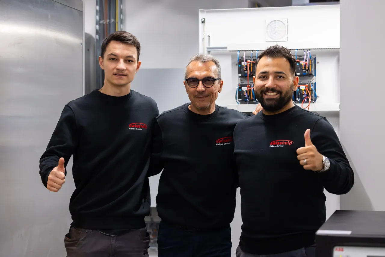 Drei Elektriker von Swisshelp Arm in Arm mit Daumen hoch vor einem Schaltschrank